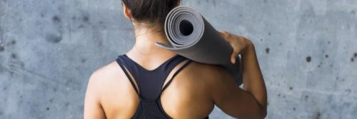 woman stands facing backward carrying yoga mat