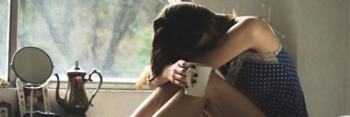 woman sits alone head on knees holding white mug feeling sad tired exhausted