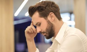 man sits in library hand in forehead thinking looking at laptop