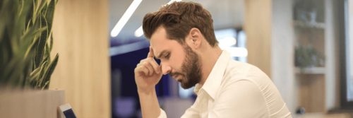 man sits in library hand in forehead thinking looking at laptop