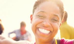 short hair woman happily smiles taking selfie on busy beach in sunny sky