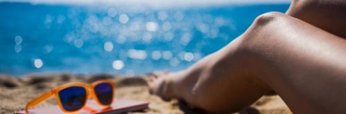 legs sunglasses book on sand beside blue ocean