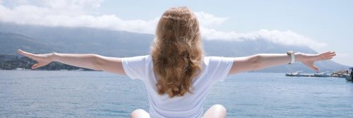 woman sits beside blue ocean facing backward gratitude life in cloudy sky