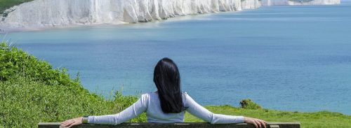 woman facing backward sits on bench looking at blue ocean