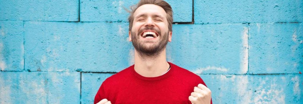 man stands holding hands closing eyes excitedly laughing shouting beside blue stone wall