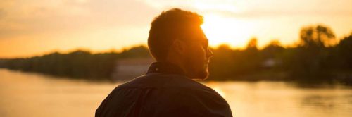 man wearing sunglasses facing backward stands beside ocean looking at sunset