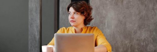 woman wearing yellow long sleeved shirt sits working on laptop looking aside