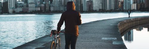 man walks his bike along footpath between ocean approaching busy city