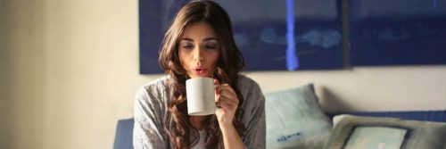 woman sits on couch holding white mug blowing