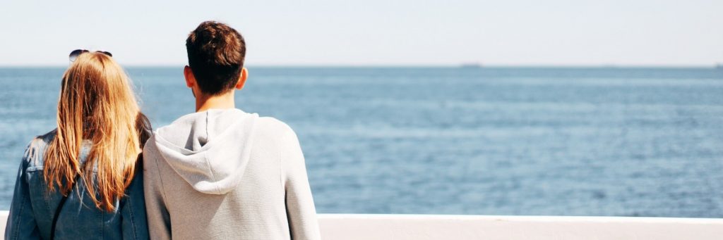 one woman one man facing backward stands on cliff watching blue ocean