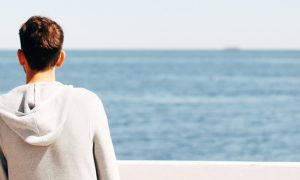 one woman one man facing backward stands on cliff watching blue ocean