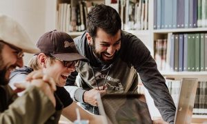 three men sits in library excitedly laughing while using laptop beside bookshelf