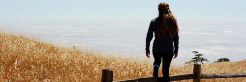 woman stands alone near cliff facing backward looking at cloudy sky