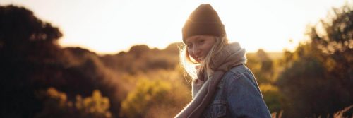 woman wearing black hat purple scarf stands in field happily smiling in sunny sky