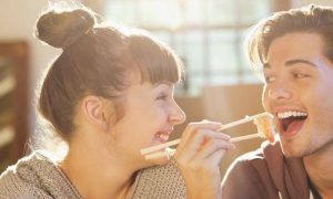 man sits using chopstick eating prawn sushi while girlfriend sitting beside laughing looking