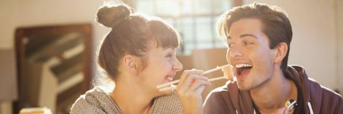 man sits using chopstick eating prawn sushi while girlfriend sitting beside laughing looking