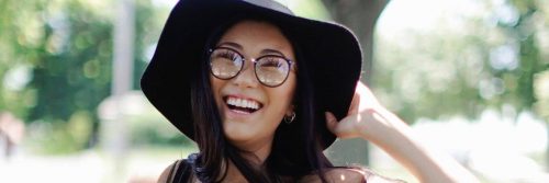 woman wearing eyelash happily smiles stands holding black hat in park