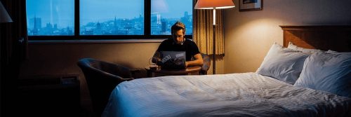 man sits beside bed in bedroom working on laptop on side table at late night