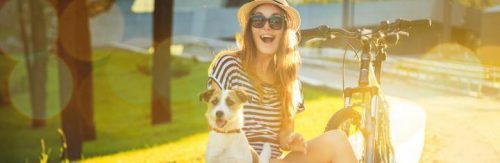 happy woman sits with little pet puppy beside bicycle in park in sunny sky