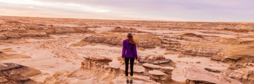 woman facing backward stands on rock in desert in cloudy sky