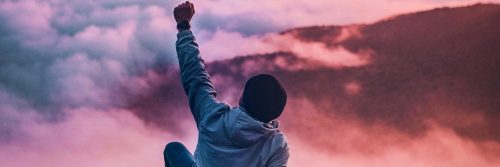 man sits raising hand looking mountain red blue cloudy sky