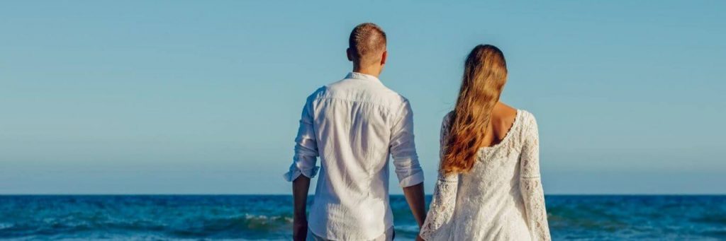 couple stands facing backward holding hands in front of blue ocean in blue clear sky