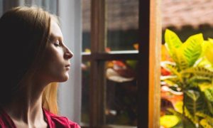 woman sits beside open window eyes closed thinking