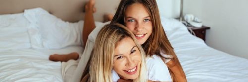 woman lies upside down on bed smiling while daughter lying on mom smiling