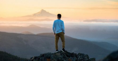 man facing backward stands on rock hands in pocket looking at mountains