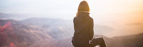 woman facing backward sits on mountain top looking at sunny sky