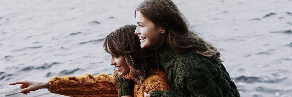 two women stands on snow hugging happily smiling pointing