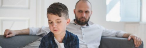 son feels sad while father sitting on couch behind angrily looking at him