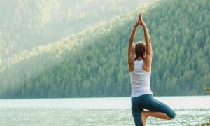 woman stands practicing yoga near lake in beautiful sunny sky