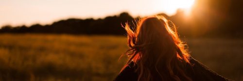 woman facing backward walks on field in sunny sky