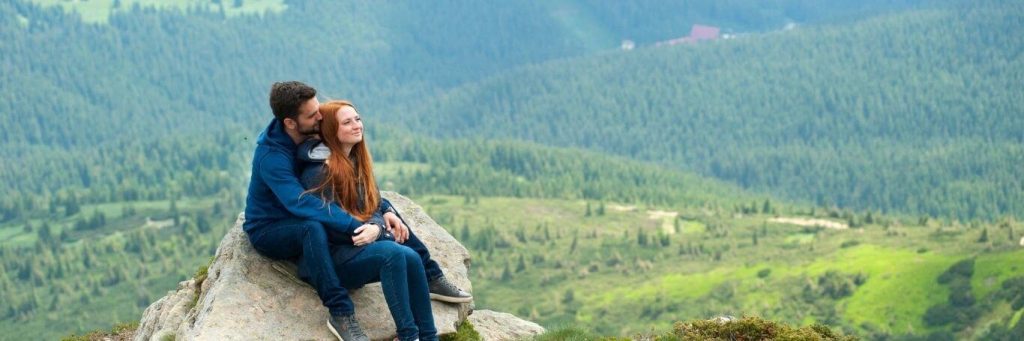 couple sits on rock while man hugging girlfriend from behind in mountainous area