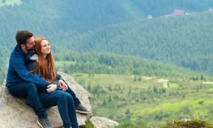 couple sits on rock while man hugging girlfriend from behind in mountainous area