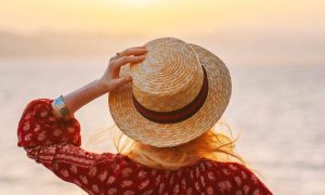 woman stands on beach hand on hat facing backward watching awesome sunset