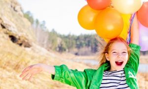 girl stands on field beside lake holding balloons happily smiling