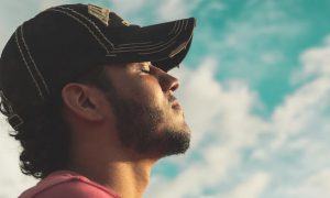 man eyes closed wears black cap meditating in blue cloudy sky