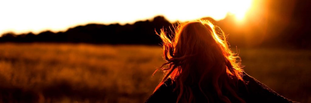 woman stands facing backward walking along field in sunny sky