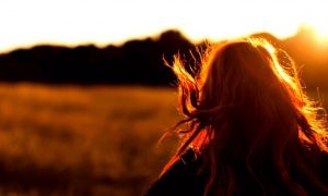 woman stands facing backward walking along field in sunny sky