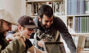 three men sits beside bookshelf in office excitedly happily looking at laptop