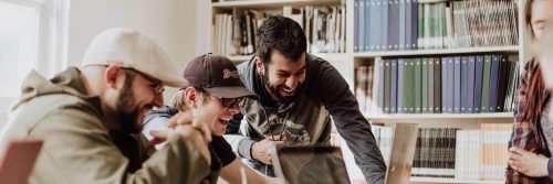 three men sits beside bookshelf in office excitedly happily looking at laptop