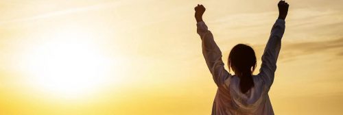 woman stands facing backward raising hands gratitude life in sunset sky