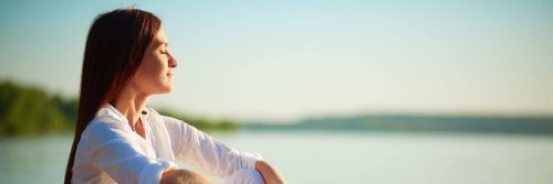 woman eyes closed sits beside lake breathing focusing on inner peace