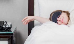 woman wearing mask covering eyes sleep on white bed set beside side table lamp telephone