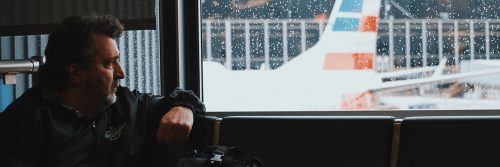 man sits on couch beside window in waiting room watching plane through glass window