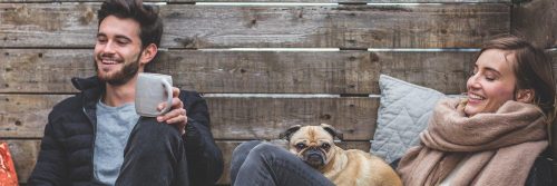 man sits beside wall holding grey mug smiling while woman sitting smiling at puppy