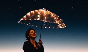 elderly woman stands holding umbrella filled up with lights happily smiling in dark star sky