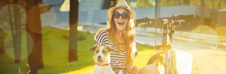 happy woman sits with little pet puppy beside bicycle in park in sunny sky